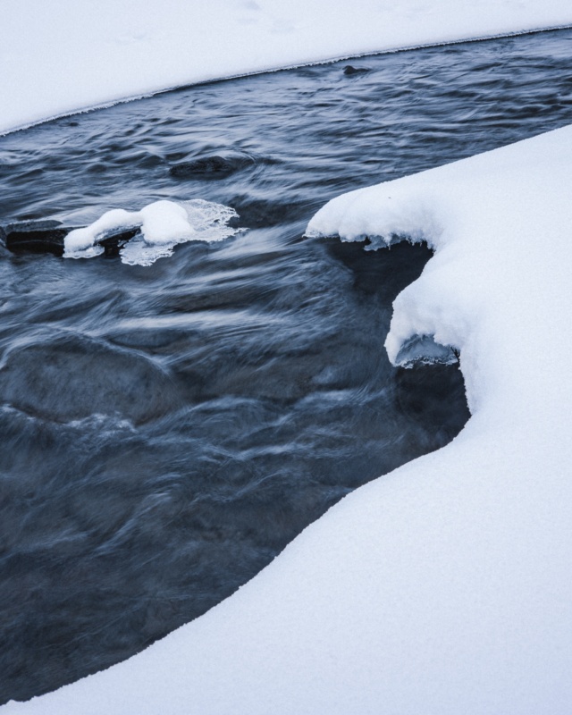 the photo depicts a stream of water rushing between snow banks.